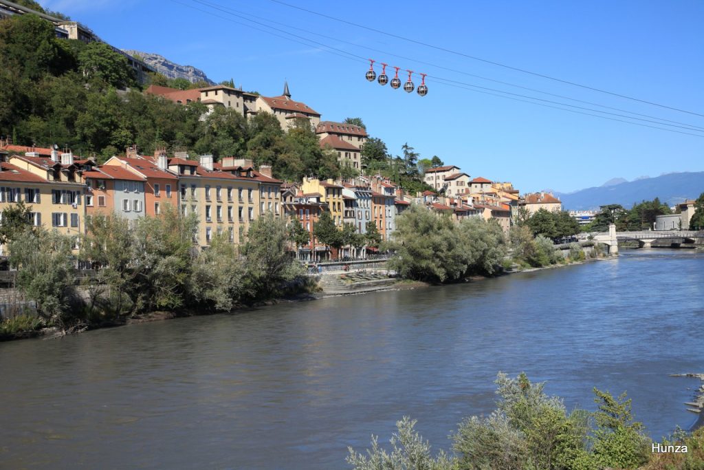 Vue sur les quais de l’Isère à Grenoble avec les maisons colorées du quartier Saint-Laurent, le téléphérique de la Bastille en arrière-plan et les montagnes des Alpes.